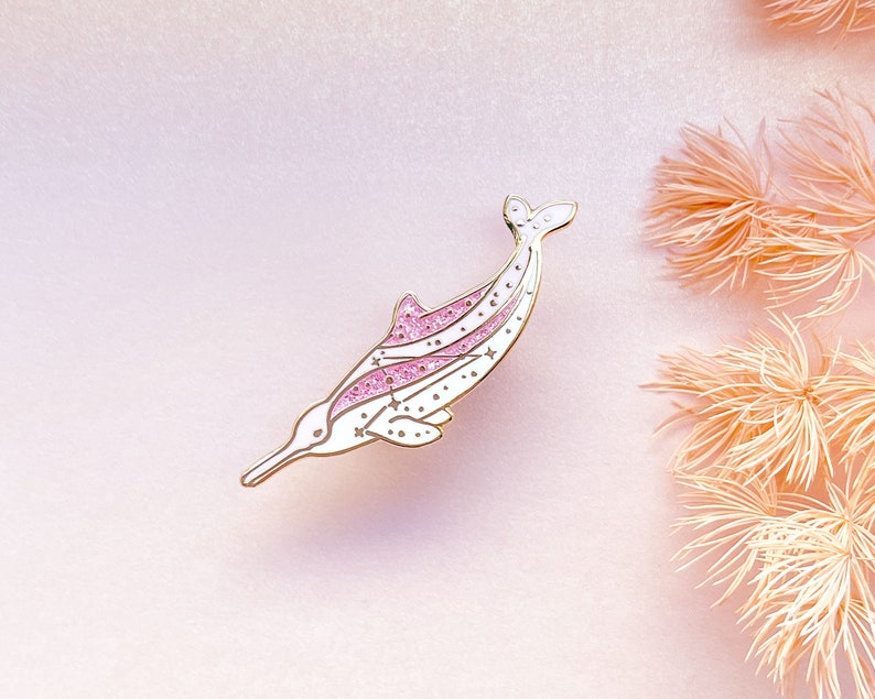 Op de afbeelding: Een wit en goudkleurige dolfijnspeld met roze glitteraccenten. De speld heeft een lange snuit, een gebogen lichaam en een staartvin. Kleine sterdetails zijn verspreid over het lichaam van de dolfijn.