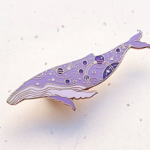 May include: A purple enamel pin shaped like a whale with a galaxy design. The whale is outlined in gold and has a white background.