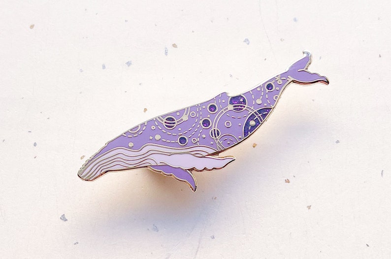 May include: A purple enamel pin shaped like a whale with a galaxy design. The whale is outlined in gold and has a white background.
