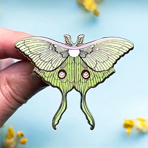 Puede incluir: Un pin de metal verde y dorado de una polilla luna con un cuerpo rosa y blanco. La polilla tiene alas y antenas detalladas.