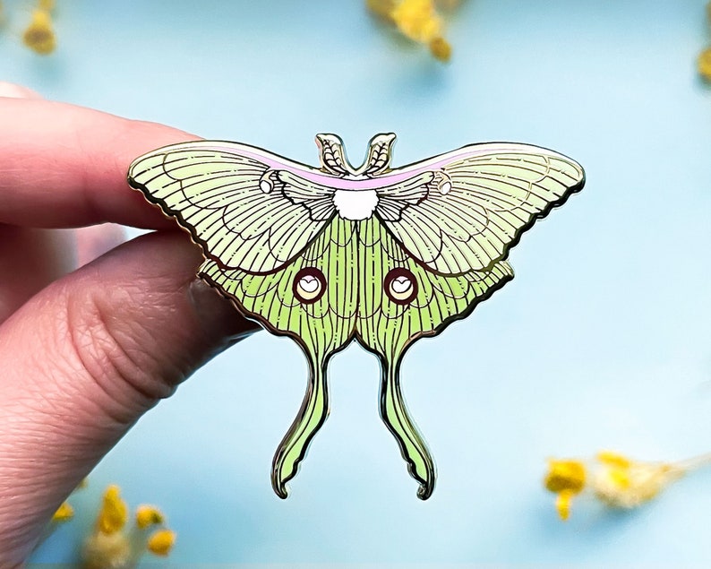 Puede incluir: Un pin de metal verde y dorado de una polilla luna con un cuerpo rosa y blanco. La polilla tiene alas y antenas detalladas.