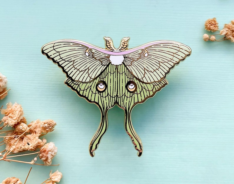 Puede incluir: Un pin de esmalte dorado con un dise&ntilde;o de polilla luna verde con detalles en rosa. La polilla tiene alas detalladas y un cuerpo delicado.