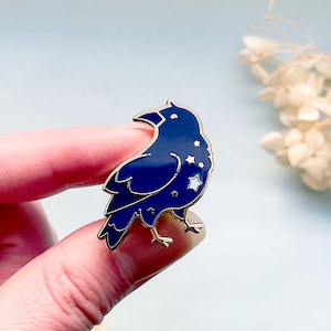 May include: A dark blue raven enamel pin with gold-colored accents. The raven features a starry design on its body and is set against a light blue background. The pin is a decorative accessory.