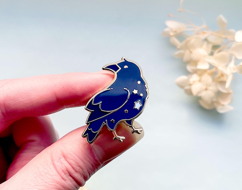 May include: A dark blue raven enamel pin with gold-colored accents. The raven features a starry design on its body and is set against a light blue background. The pin is a decorative accessory.