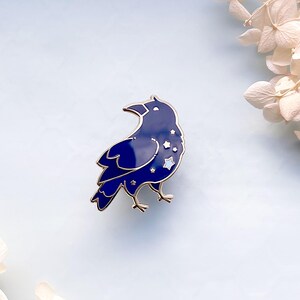 May include: A navy blue enamel pin in the shape of a raven, with gold-colored trim. The raven's body features a starry night design with white stars. The pin is a decorative accessory.