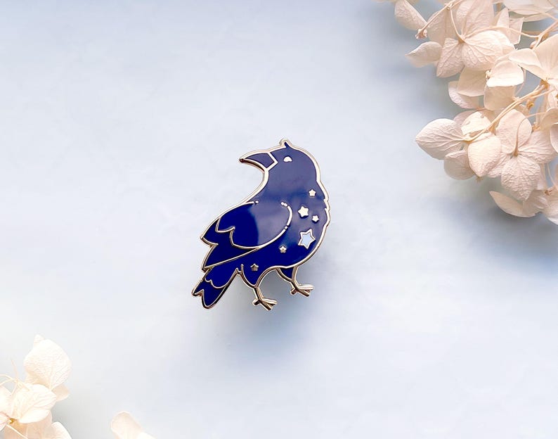 May include: A navy blue enamel pin in the shape of a raven, with gold-colored trim. The raven's body features a starry night design with white stars. The pin is a decorative accessory.