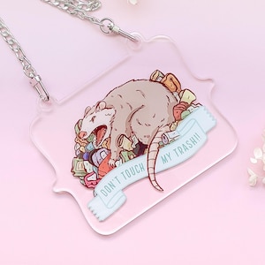 May include: A clear acrylic keychain with a cartoon illustration of a possum surrounded by trash. The possum is brown and white and has a pink nose. The text on the keychain reads "Don't touch my trash!"