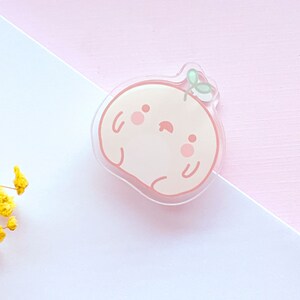 May include: A peach-colored cartoon character with a small green leaf on top. The character has a cute, smiling face with closed eyes and rosy cheeks. The character is made of clear acrylic.