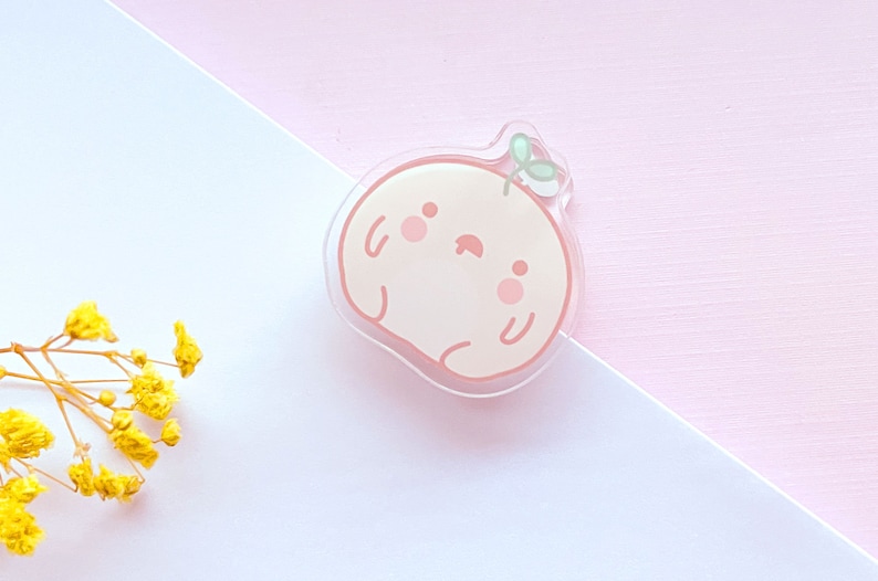 May include: A peach-colored cartoon character with a small green leaf on top. The character has a cute, smiling face with closed eyes and rosy cheeks. The character is made of clear acrylic.