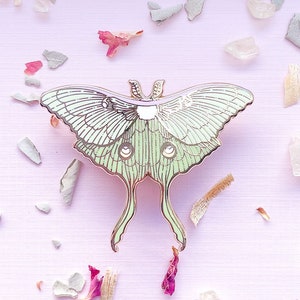 Puede incluir: Un pin de metal verde y dorado de una polilla luna con alas y antenas detalladas. El pin est&aacute; sobre un fondo rosa con p&eacute;talos de flores esparcidos.