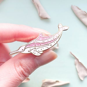 Op de afbeelding: Een goudkleurige emaille pin in de vorm van een roze dolfijn met witte accenten. De dolfijn heeft een glitterende roze streep en ster details. De pin wordt tegen een lichtblauwe achtergrond gehouden.