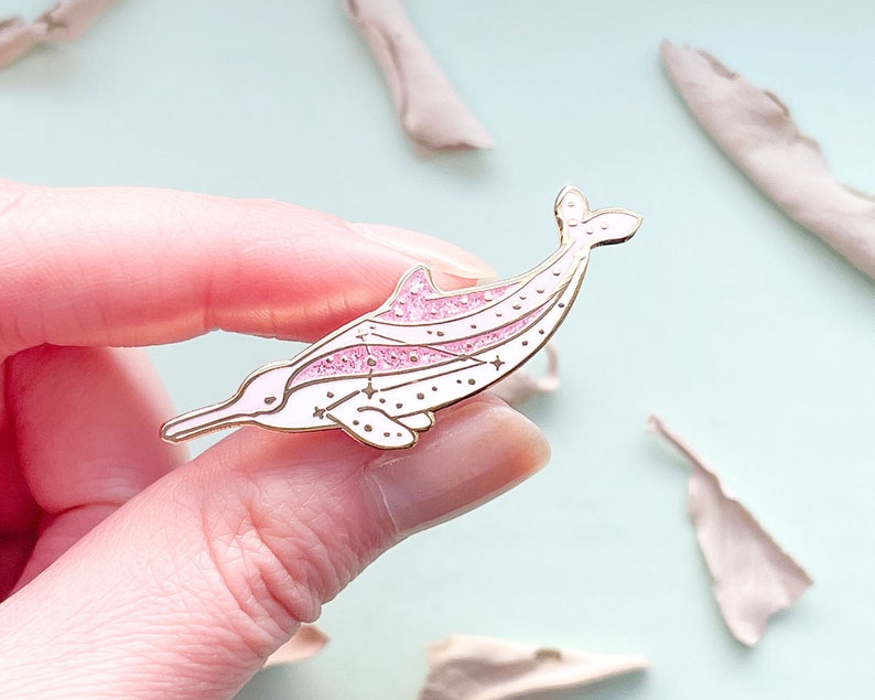 Op de afbeelding: Een goudkleurige emaille pin in de vorm van een roze dolfijn met witte accenten. De dolfijn heeft een glitterende roze streep en ster details. De pin wordt tegen een lichtblauwe achtergrond gehouden.