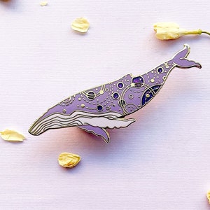 May include: A purple and gold enamel pin of a whale with a galaxy pattern. The whale is outlined in gold and has a white underbelly.
