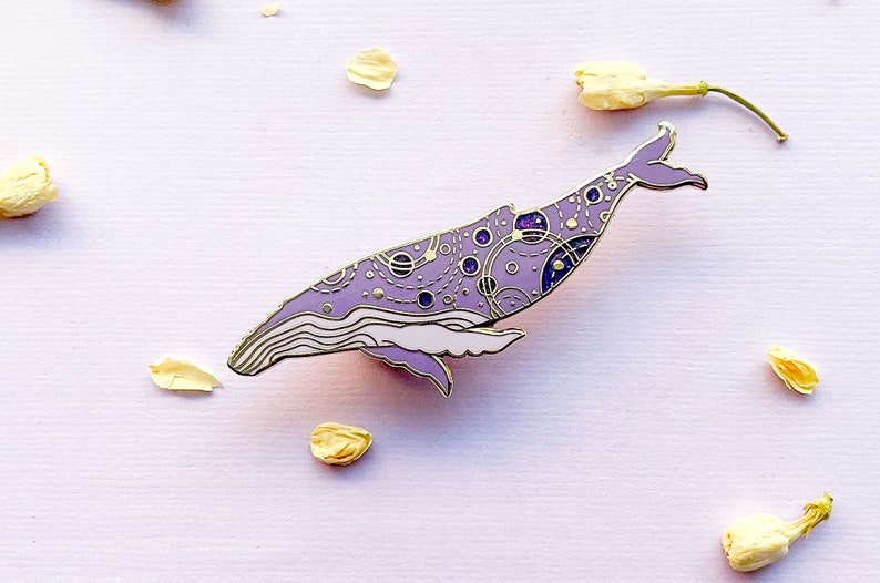 May include: A purple and gold enamel pin of a whale with a galaxy pattern. The whale is outlined in gold and has a white underbelly.