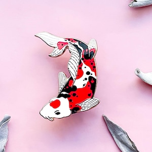 Puede incluir: Pin decorativo de carpa koi con detalles esmaltados en rojo, negro y blanco. El pin presenta un diseño detallado del pez, con un contorno metálico brillante. El pin está sobre un fondo rosa con elementos decorativos.