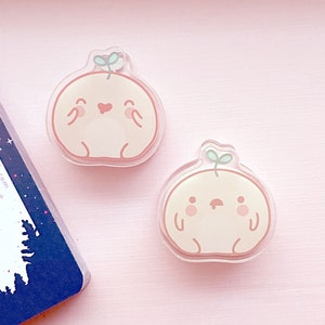May include: Two peach-colored cartoon characters with a small green sprout on top. The characters have a round body, small arms and legs, and a smiling face. The characters are made of clear acrylic and are about 2 inches in diameter.