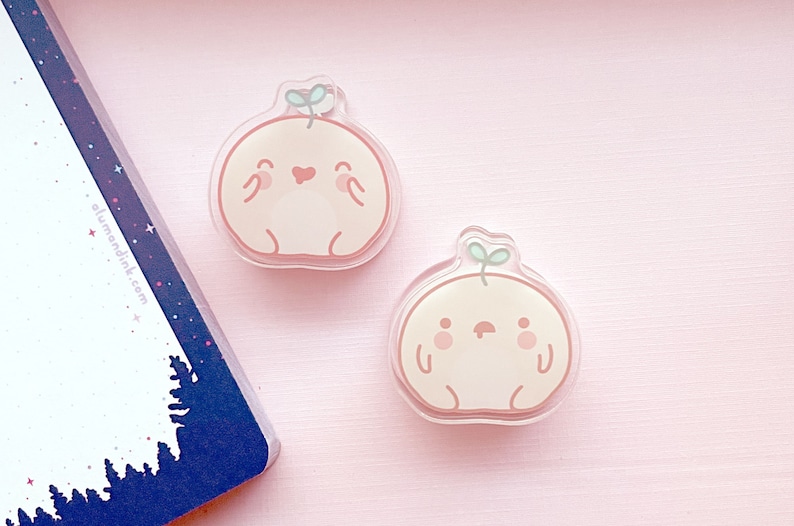 May include: Two peach-colored cartoon characters with a small green sprout on top. The characters have a round body, small arms and legs, and a smiling face. The characters are made of clear acrylic and are about 2 inches in diameter.
