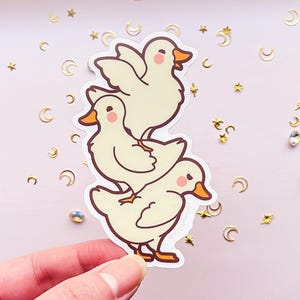 May include: A sticker featuring three cartoon ducks stacked on top of each other. The ducks are white with orange beaks and feet. The sticker is on a pink background with gold glitter.