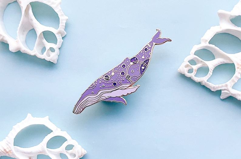 May include: Purple and gold enamel pin of a whale with a detailed pattern of circles and lines. The whale is facing left and has a slightly upturned tail.