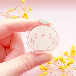 May include: A clear pink and white acrylic charm with a cartoon character design. The character is a round, white creature with a pink face, small eyes, and a small green sprout on top of its head.