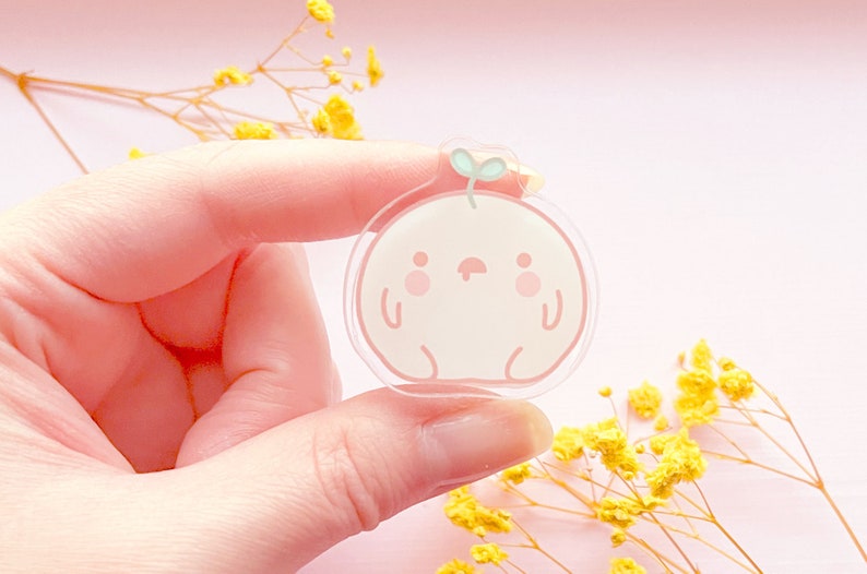 May include: A clear pink and white acrylic charm with a cartoon character design. The character is a round, white creature with a pink face, small eyes, and a small green sprout on top of its head.