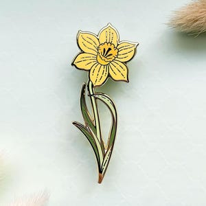 May include: A daffodil flower pin with a yellow bloom and green stem. The pin features a detailed design with black outlines and a gold-colored metal finish. The flower is approximately 2 inches tall.