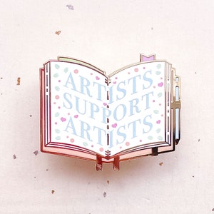May include: Gold-tone enamel pin shaped like an open book with the words "Artists Support Artists" in blue and pink lettering on a white background. The book has a pink and green floral design.