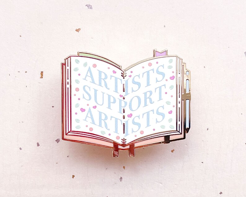 May include: Gold-tone enamel pin shaped like an open book with the words "Artists Support Artists" in blue and pink lettering on a white background. The book has a pink and green floral design.