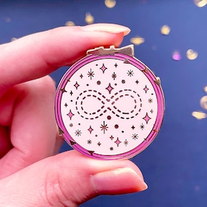 May include: Gold-tone pin with a pink enamel design. The design features an infinity symbol surrounded by stars and dots.