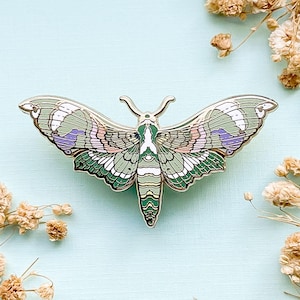 May include: A detailed metal pin in the shape of a moth, featuring intricate wing patterns in shades of green, white, pink, and purple. The pin has a silver-colored outline and antenna. The moth pin is a decorative accessory.