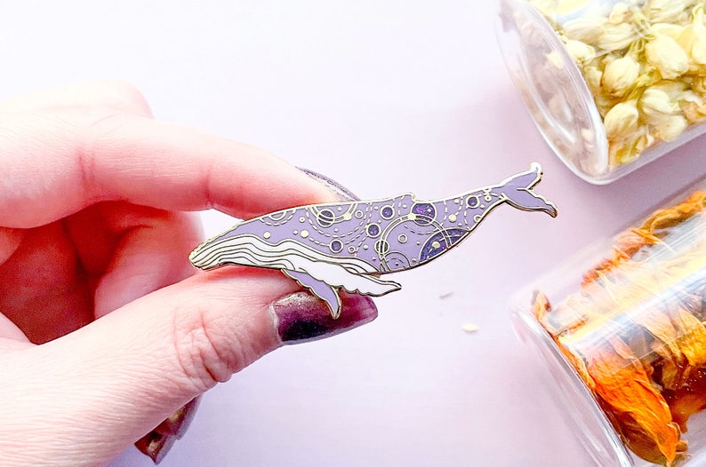 May include: A purple enamel pin of a whale with a galaxy pattern. The whale is outlined in gold and has white details. The pin is a great addition to any whale lover's collection.