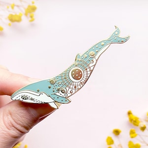 May include: A gold-tone enamel pin featuring a blue whale with a celestial design. The whale is outlined in gold and has a white belly. The celestial design includes stars, moons, and planets.