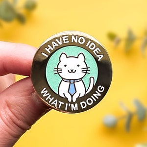 Puede incluir: Pin de color dorado con un gato blanco que lleva una corbata azul. El pin dice "No tengo ni idea de lo que estoy haciendo".