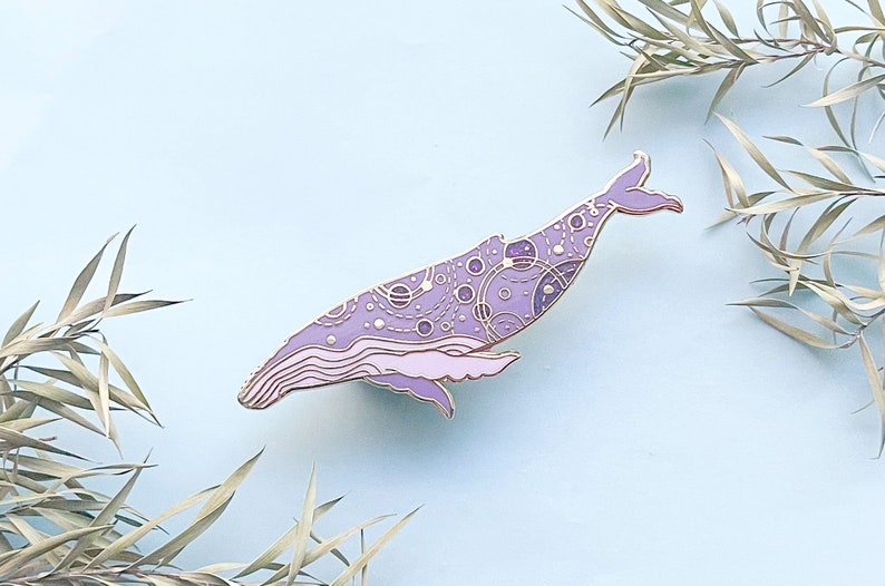 May include: A purple and gold enamel pin of a whale with a galaxy pattern. The whale is outlined in gold and has a detailed design of planets and stars.