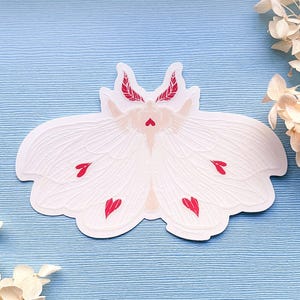 May include: White paper sticker of a moth with red hearts on its wings. The moth has a delicate, intricate design.