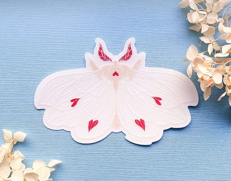 May include: White paper sticker of a moth with red hearts on its wings. The moth has a delicate, intricate design.