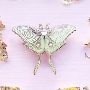 Puede incluir: Un pin de metal verde y dorado de una polilla luna con alas y antenas detalladas.