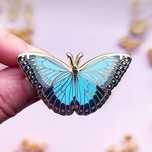 May include: A blue and black butterfly enamel pin with gold accents. The butterfly has a light blue body and black wings with white spots.