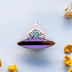 May include: A purple and silver enamel pin of a blue alien in a flying saucer. The alien is smiling and has its arms raised.