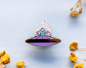 May include: A purple and silver enamel pin of a blue alien in a flying saucer. The alien is smiling and has its arms raised.