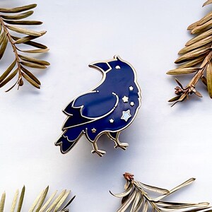 May include: A navy blue raven enamel pin with gold accents and star details. The raven is surrounded by gold metal and has a glossy finish. The pin is a decorative accessory.