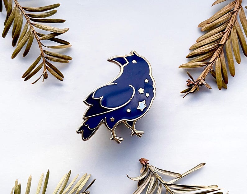 May include: A navy blue raven enamel pin with gold accents and star details. The raven is surrounded by gold metal and has a glossy finish. The pin is a decorative accessory.