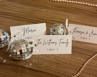 May include: Wedding table setting with place cards and disco ball decorations. The place cards are white with black calligraphy names and table numbers. Small disco balls are attached with white ribbons. A string of pearls is in the background.
