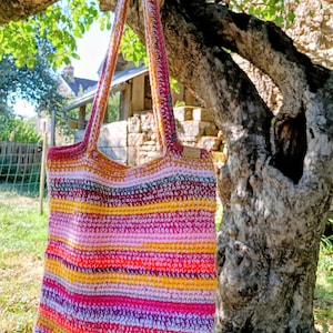 Pode incluir: Uma bolsa tote de crochê feita à mão com listras horizontais em tons vibrantes de rosa, laranja, amarelo e roxo. A bolsa possui duas alças longas de ombro e uma pequena etiqueta retangular fixada no lado superior direito do corpo.