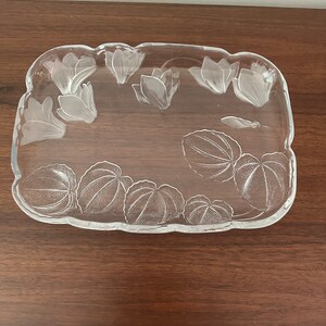 May include: A clear glass serving tray with a rectangular shape and scalloped edges. The tray features etched floral designs, including stylized flowers and leaves. The tray is displayed on a dark brown wooden surface.