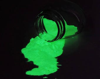 Könnte beinhalten: Ein Glas mit grünem, im Dunkeln leuchtendem Pulver kippt auf eine schwarze Oberfläche. Das Pulver leuchtet hell im Dunkeln.