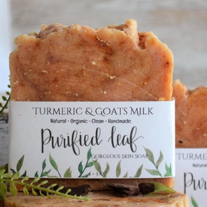 May include: Two bars of handmade soap with a label that reads "Turmeric & Goats Milk Natural Organic Clean Handmade Purified Leaf Gorgeous Skin Soap". The soap is a light orange color and has a rough texture.