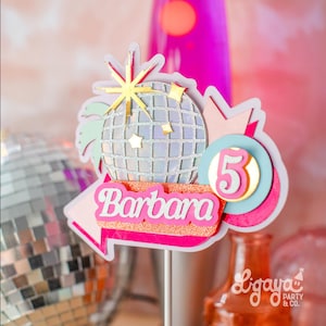 Puede incluir: Un adorno para pastel rosa y blanco con una bola de discoteca, una explosión de estrellas y el nombre "Barbara" en una flecha rosa. El número "5" está en un círculo rosa con un contorno dorado. El adorno para pastel está en un palo blanco.