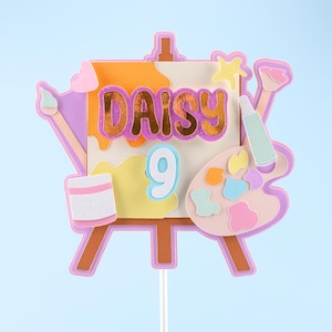 Pode incluir: Um topo de bolo colorido com um cavalete de artista com tela, paleta de pintura e pincel. O nome "DAISY" é exibido em letras douradas, com o número "9" abaixo. O design inclui tons de rosa, laranja, amarelo e roxo, perfeito para uma festa de aniversário.