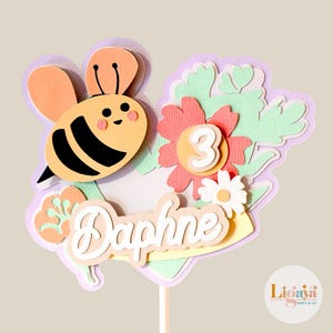 Puede incluir: Un adorno para pastel colorido con una abeja, flores, el nombre "Daphne" y el número "3". El adorno está en un palo y es perfecto para un pastel de cumpleaños.